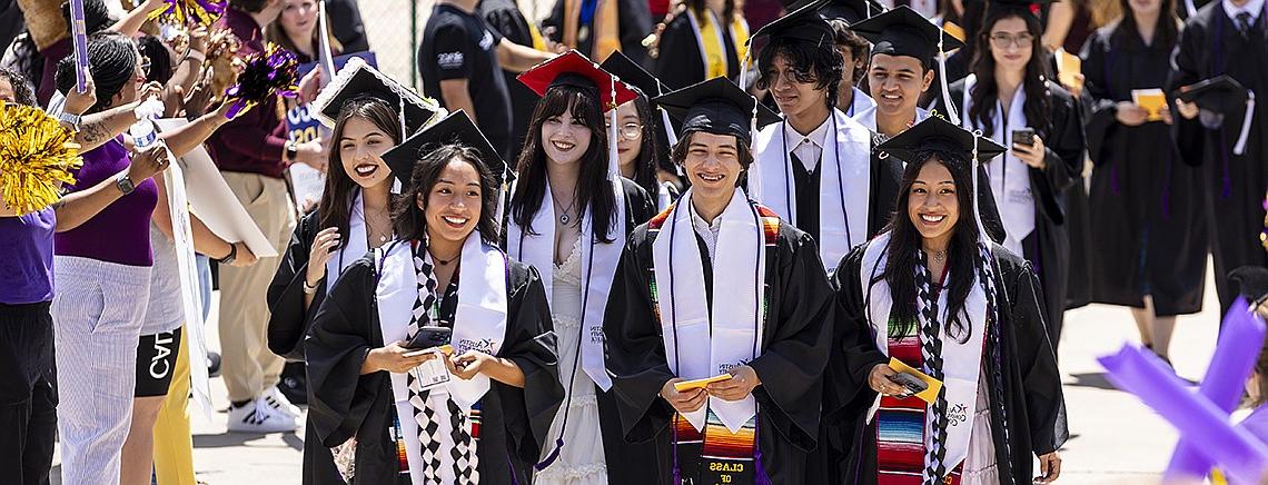 A group of smiling graduates in caps and gowns walk together at a commencement ceremony, with colorful stoles and sashes. They are surrounded by cheering supporters holding pom-poms and taking photos.